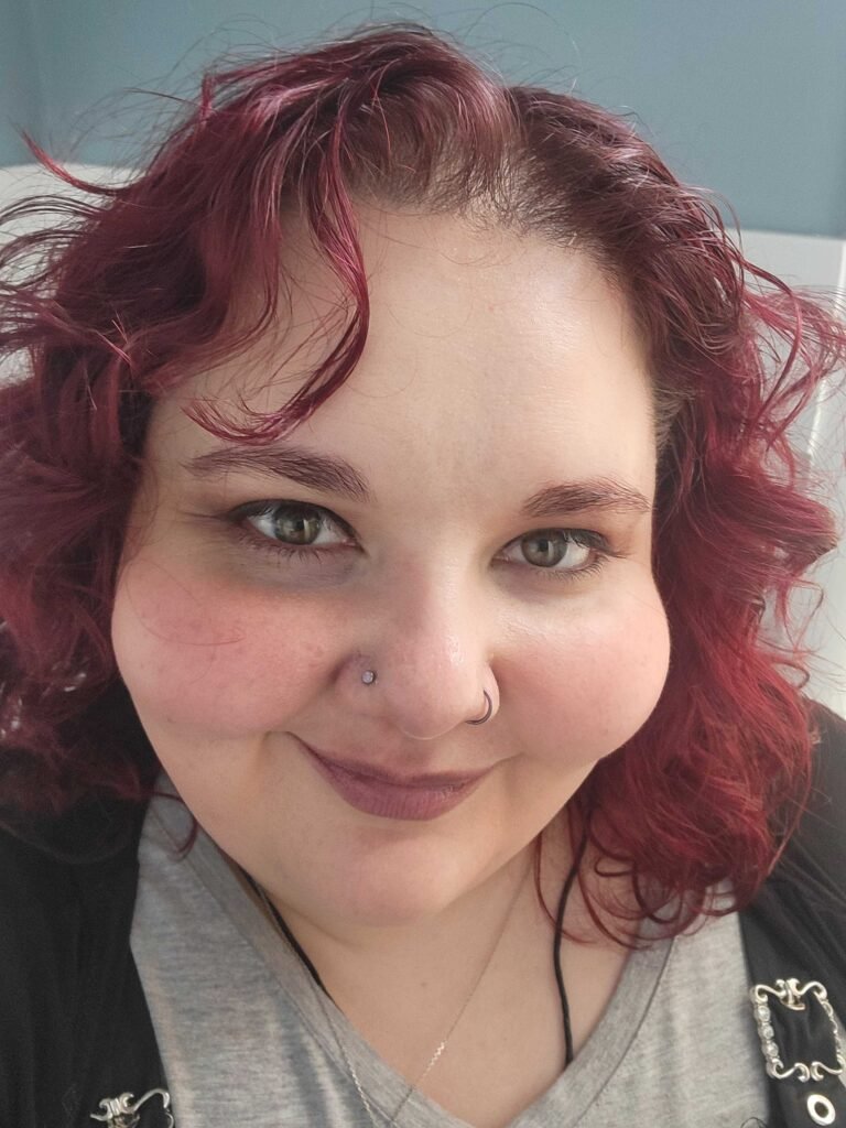 Woman smiling at camera with burgundy hair in wavy curls, two piercings on her nose, and necklaces below a grey shirt